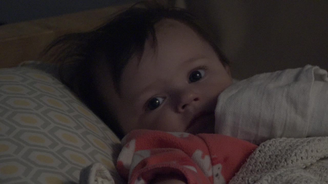 At 0:00, a baby in a pink sleeper looks toward the camera while lying beside pale blue and yellow patterned cushions in Westminster, Colorado.