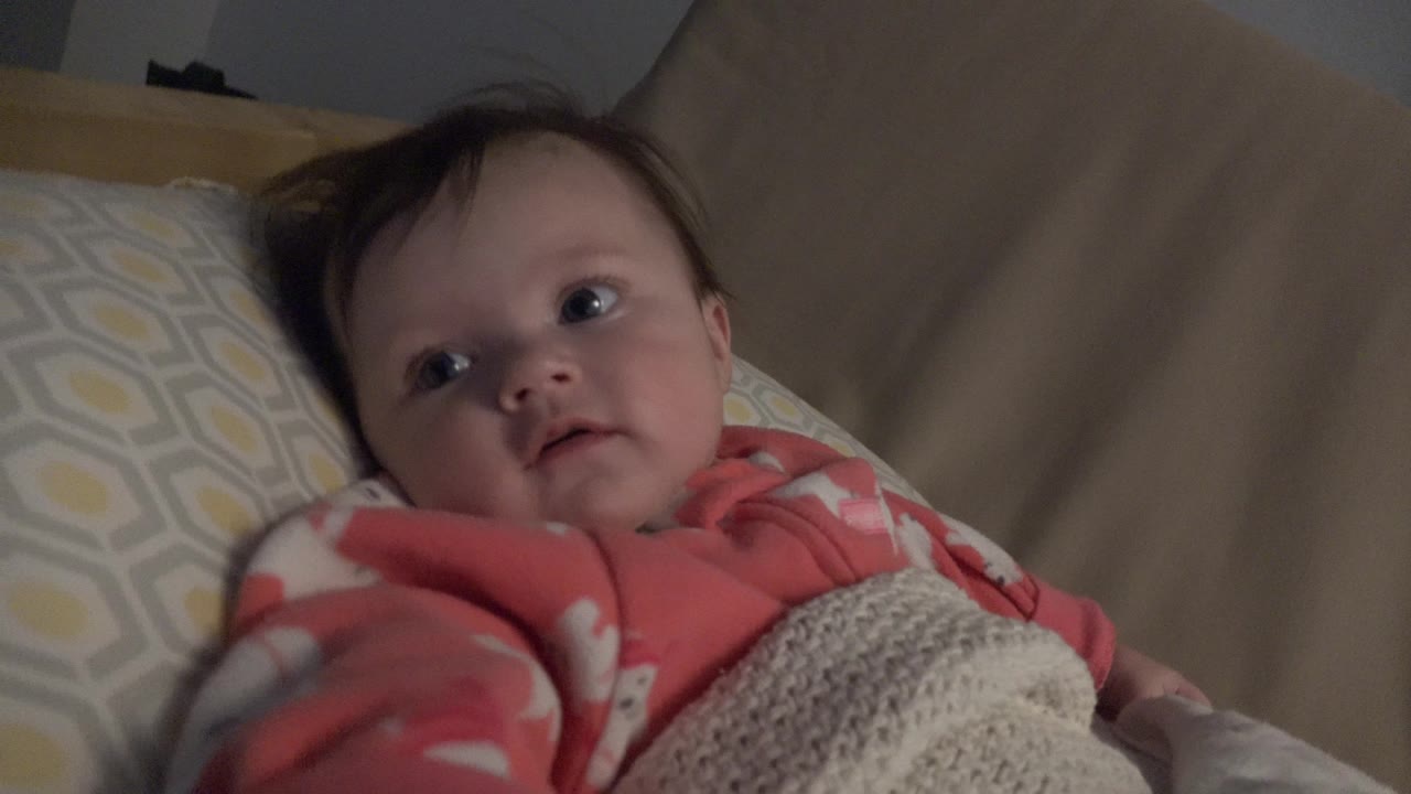 At 0:01, the baby remains tucked under the knitted blanket and looks slightly toward the side of the camera in the Westminster bedroom setting.