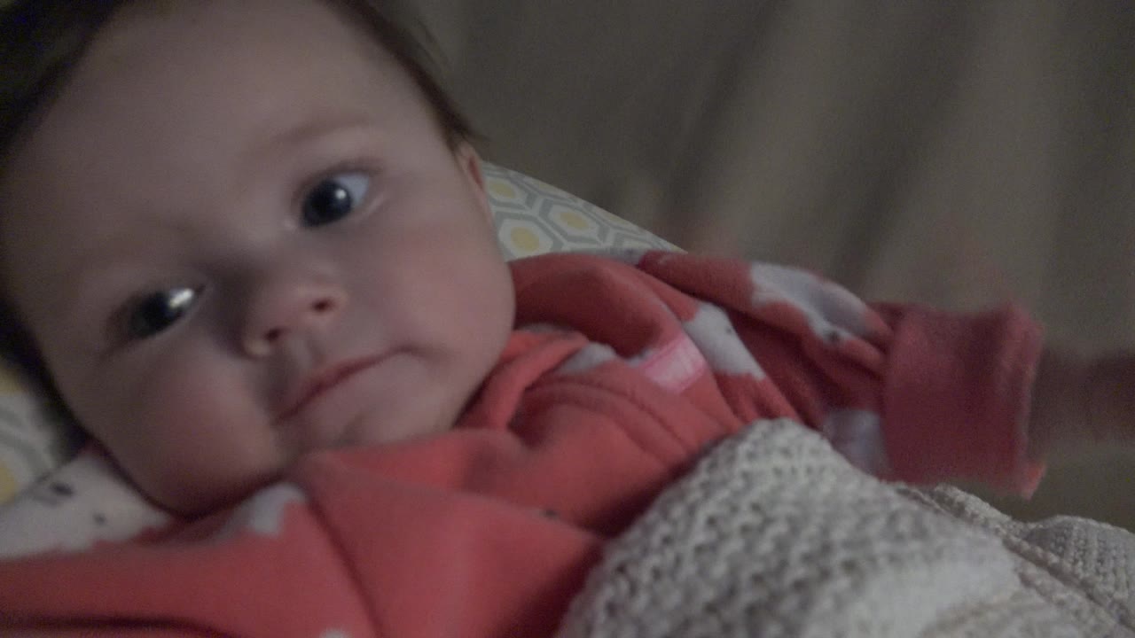 The baby remains reclined in the Westminster, Colorado crib scene, looking toward the lens while the blanket covers the lower body.