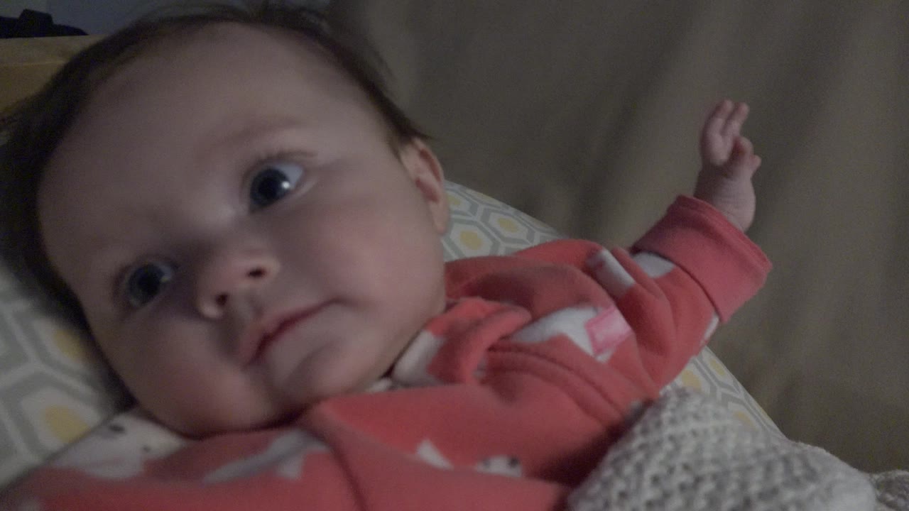 The baby remains on the blanket in Westminster, Colorado, with wide eyes turned sideways and the small raised hand visible above the shoulder.