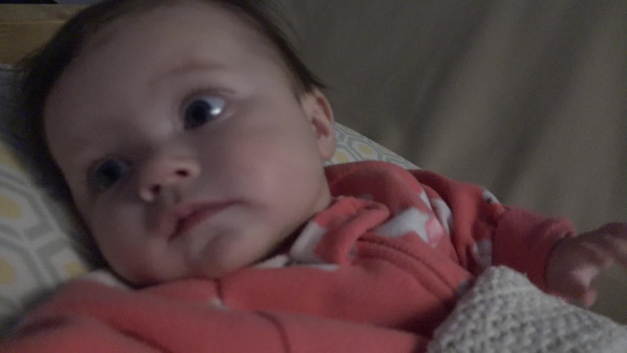 At 0:01, the baby remains reclined on the patterned bedding, with the face angled upward and the coral sleeper visible across the foreground.