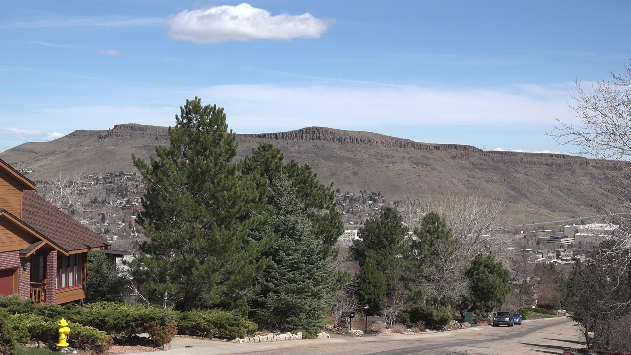 Opening frame of a Westminster, Colorado residential street with evergreen trees, a partial house at left, distant buildings, brown hills, blue sky, and no visible people.