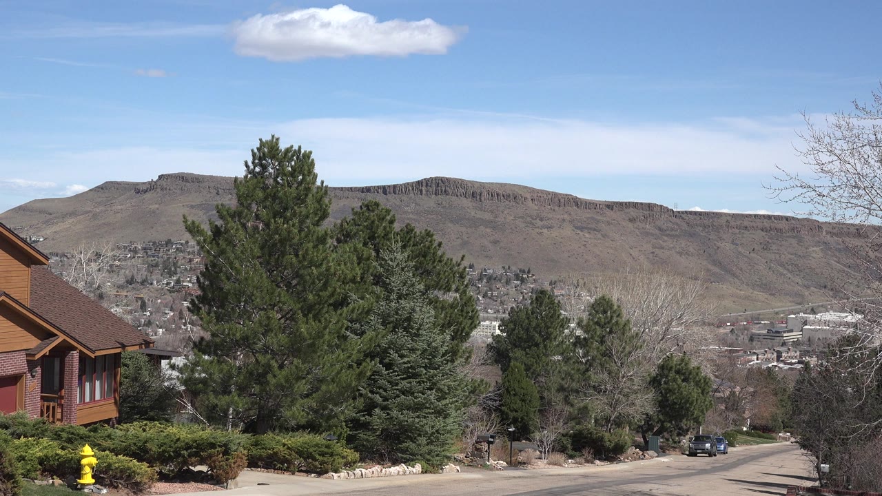 Second frame in Westminster, Colorado with a partial home on the left, evergreens around the road, distant city buildings below the ridge, and no visible people.