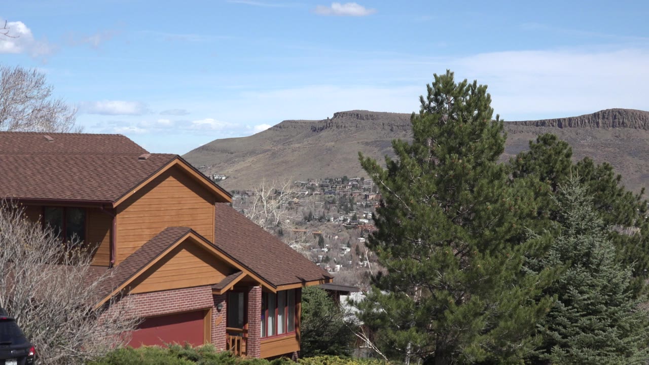 Opening frame of a Westminster, Colorado house partly screened by evergreens, with a developed valley and flat-topped hills in the distance, no visible people.