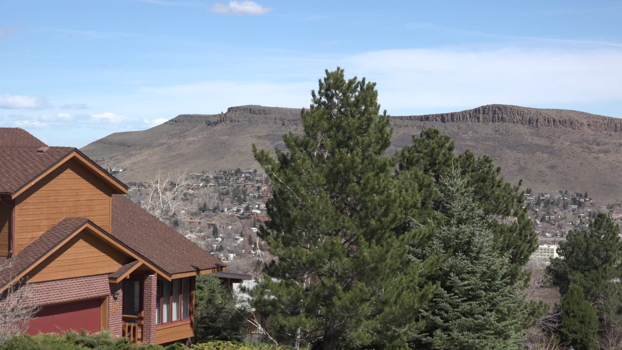 Brown house, evergreen trees, distant homes, and dry ridge under blue sky.