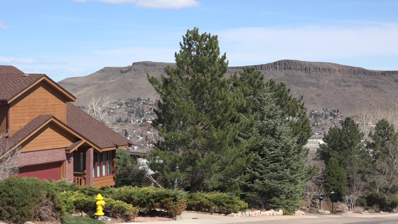 First frame of a Westminster, Colorado residential exterior with a wooden house at left, evergreens across the middle, brown hills in the distance, and no visible people.