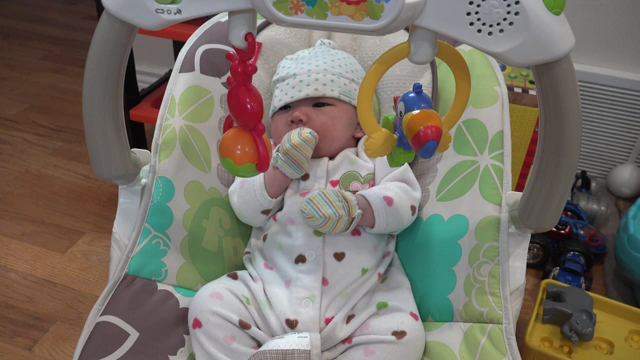 A newborn in Westminster, Colorado lies in a patterned baby carriage, wearing a white sleeper, striped mittens, and a dotted cap while red and yellow toys hang close to the face.