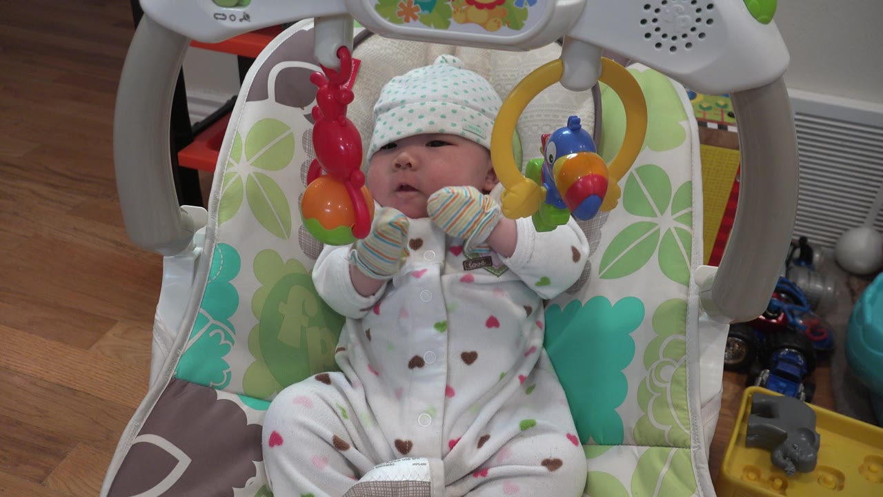 A newborn in a Westminster, Colorado baby carriage looks upward between the red toy on the left and the yellow ring toy on the right, with the room floor visible behind.