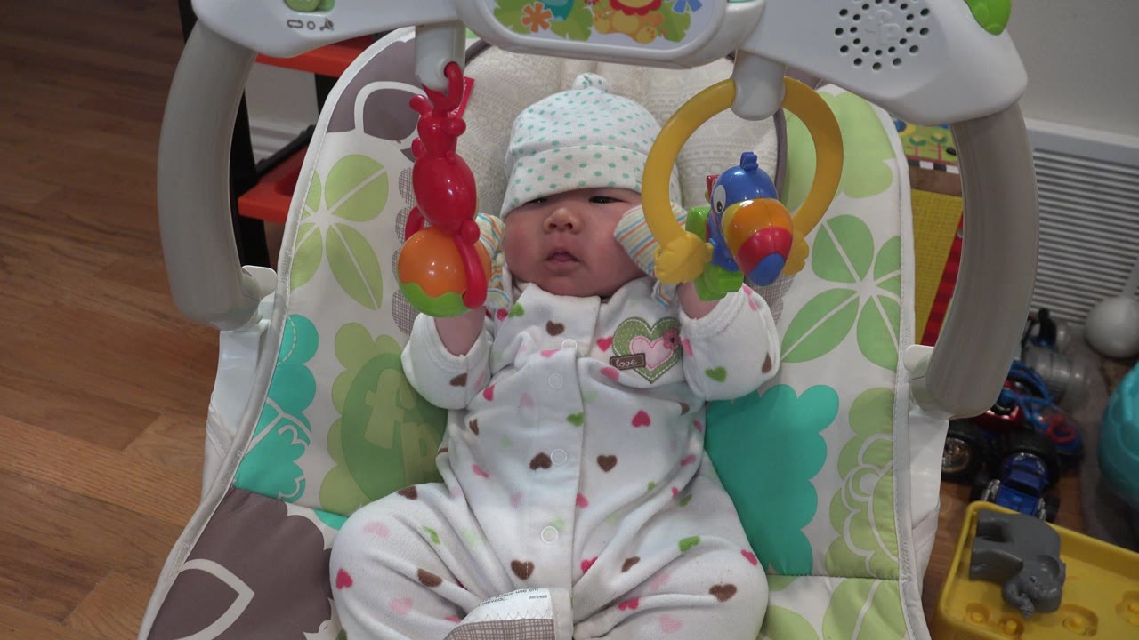Newborn in sleeper with raised arms beneath hanging toys.