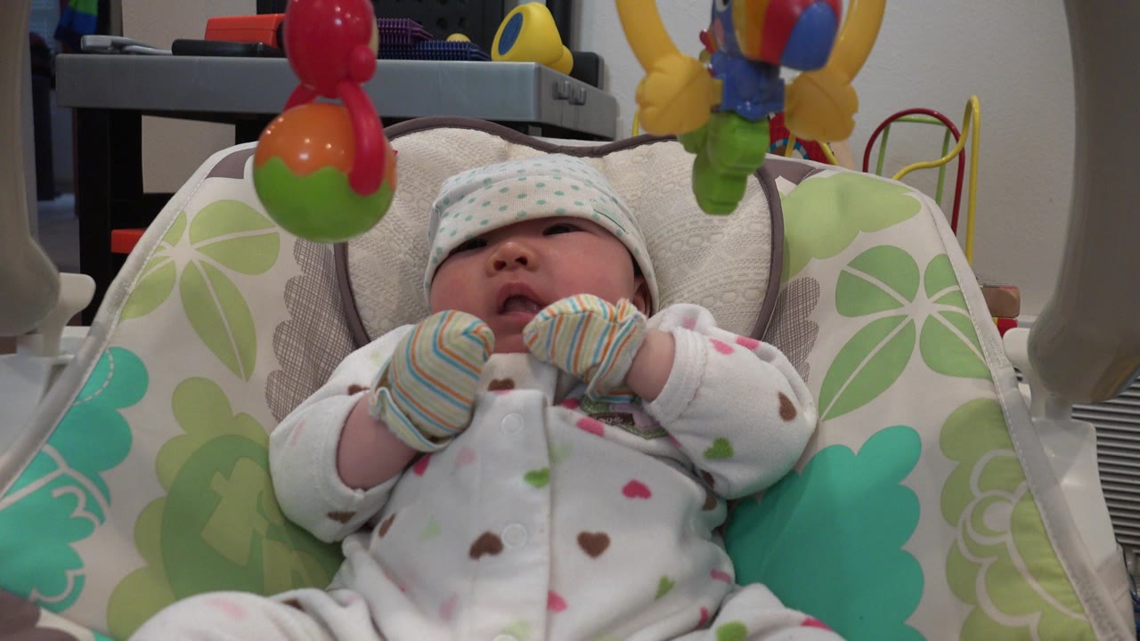 At 0:00, a young infant in Westminster, Colorado rests in a green and white baby carrier with colorful hanging toys above the face.