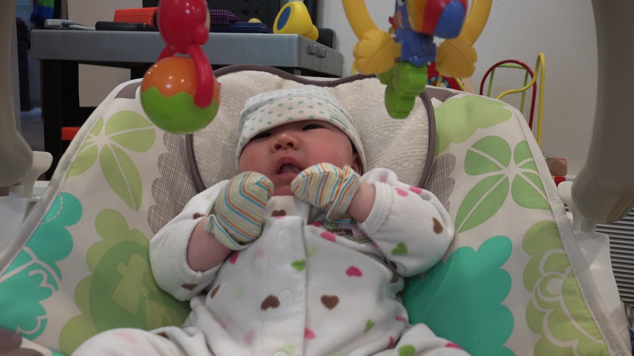 At 0:01, the infant remains centered in the Westminster indoor scene, with striped mittens held near the chest and a red hanging toy above.