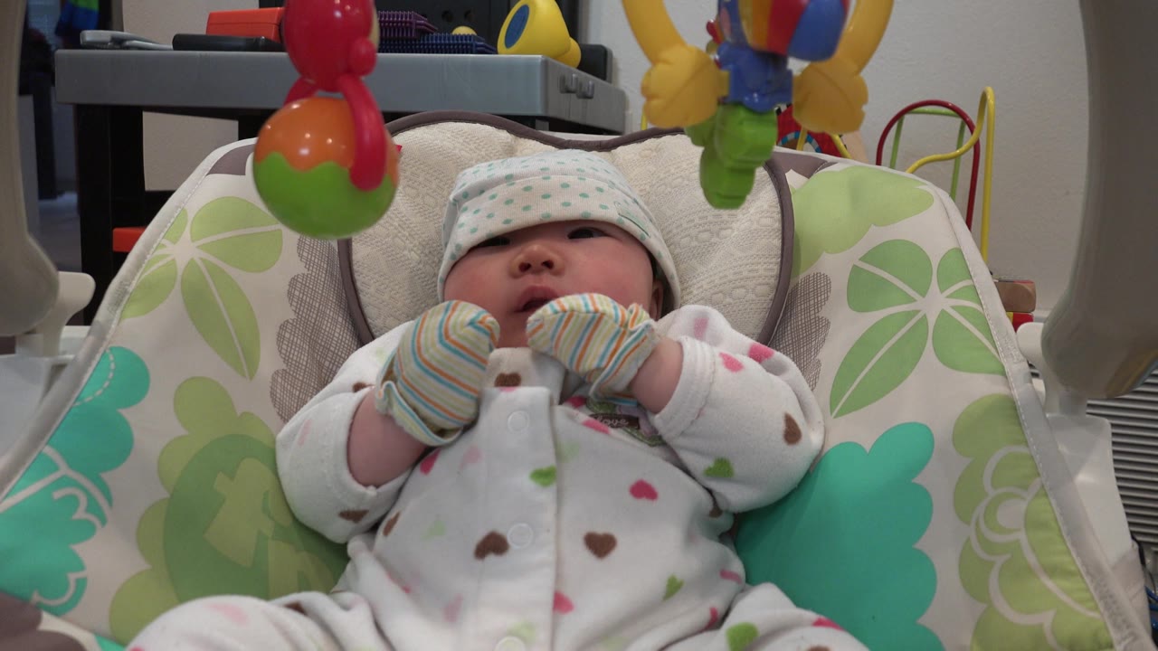Infant in mittens beneath a colorful toy bar.