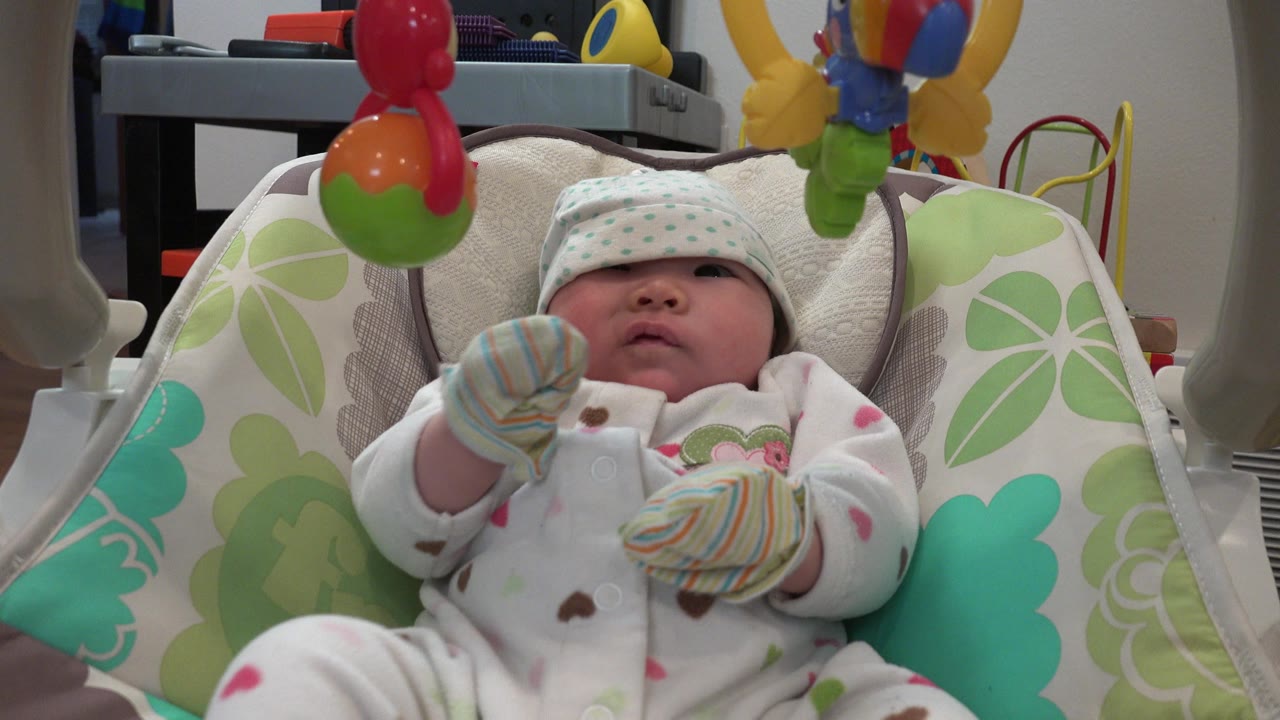 Baby lying in a cushioned infant seat beneath hanging toys.