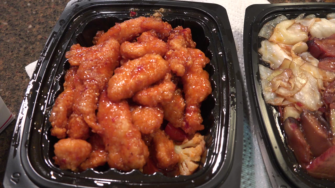 Black takeout tray filled with orange sauced fried meat beside another tray of vegetables.