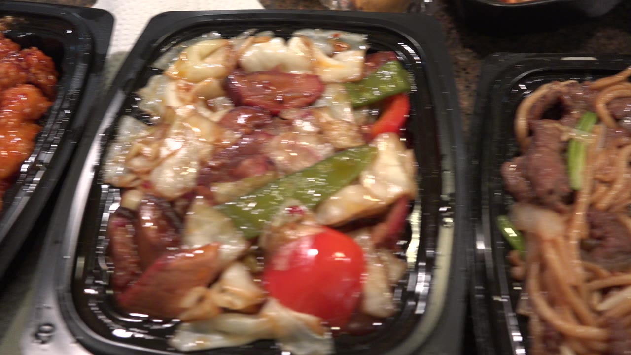 Close-up of sauced vegetables and beef in a black plastic food container beside noodles.