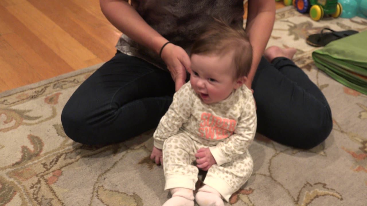 In Westminster, Colorado, a baby sits on a patterned rug while a cropped adult kneels behind with hands near the baby's shoulders.