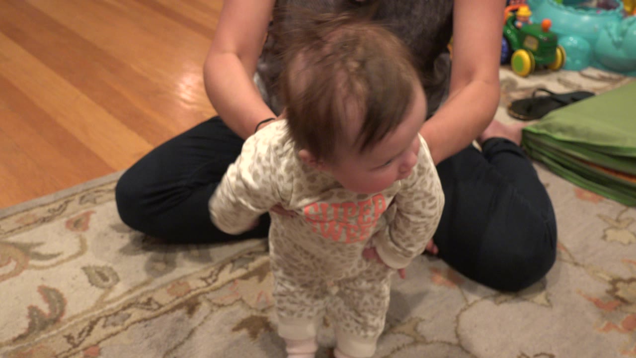 Baby standing on a rug with an adult seated behind.
