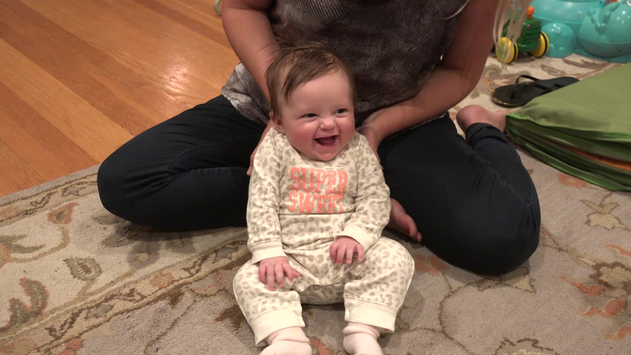 Laughing baby seated between an adult woman's legs on a rug.