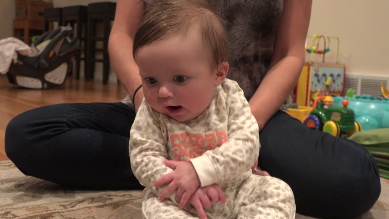 Baby sitting on a rug with mother seated behind her.