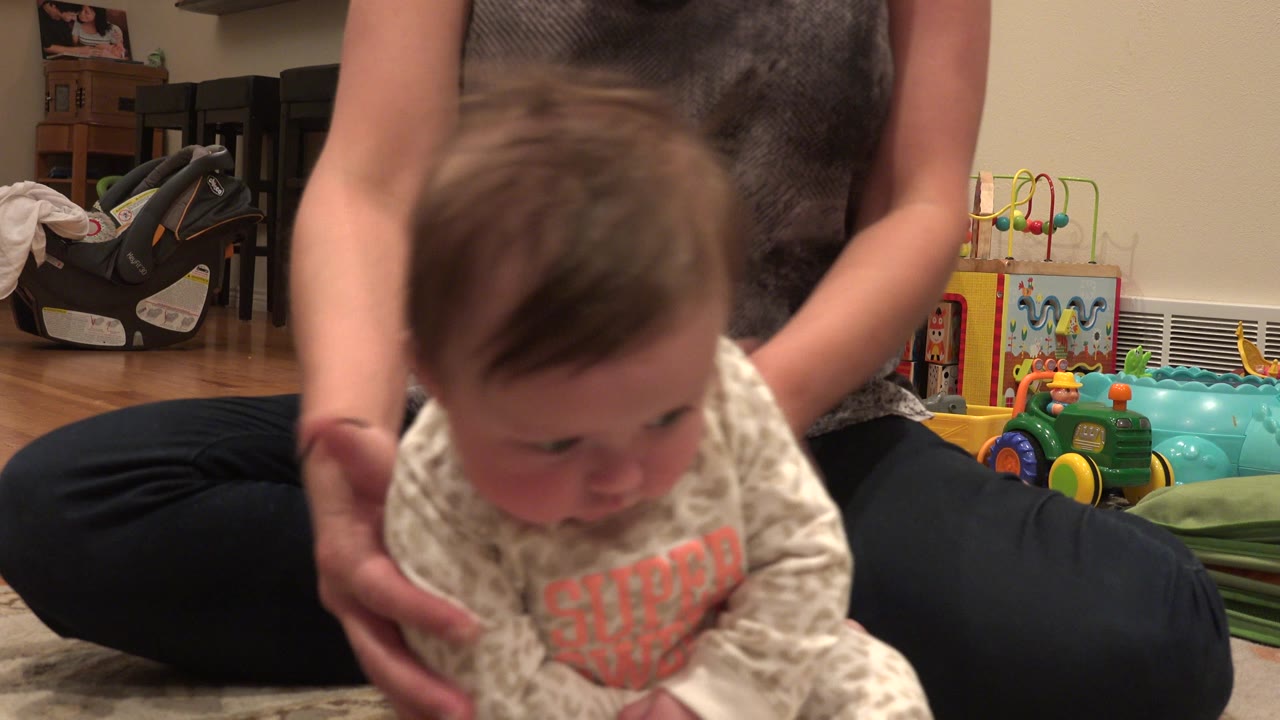 In Westminster, Colorado, a baby leans forward in a patterned sleeper while an adult's hands support them from behind on a living room rug.