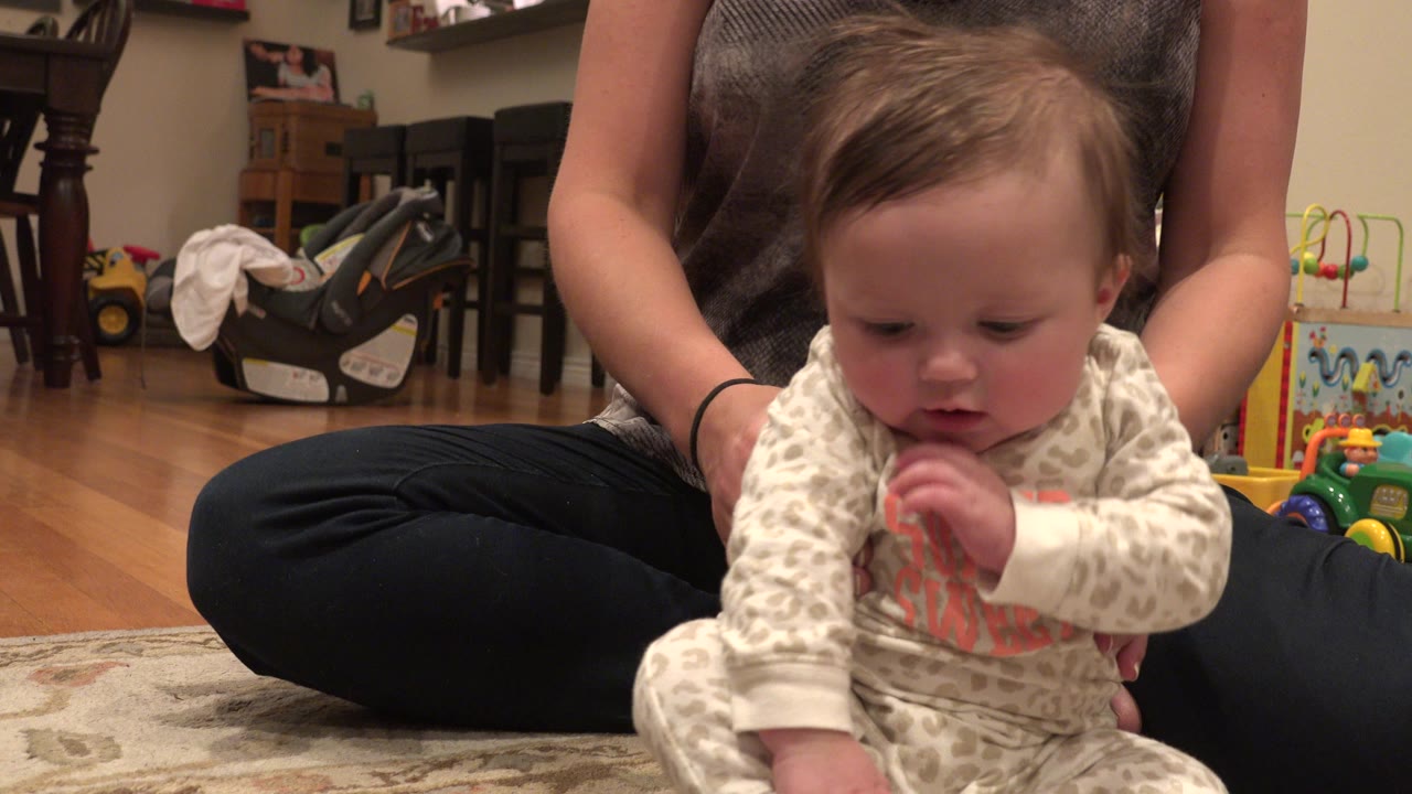 In Westminster, Colorado, a baby in a white patterned sleeper sits close to the camera on a rug while a blonde woman in black clothing holds the child from behind.