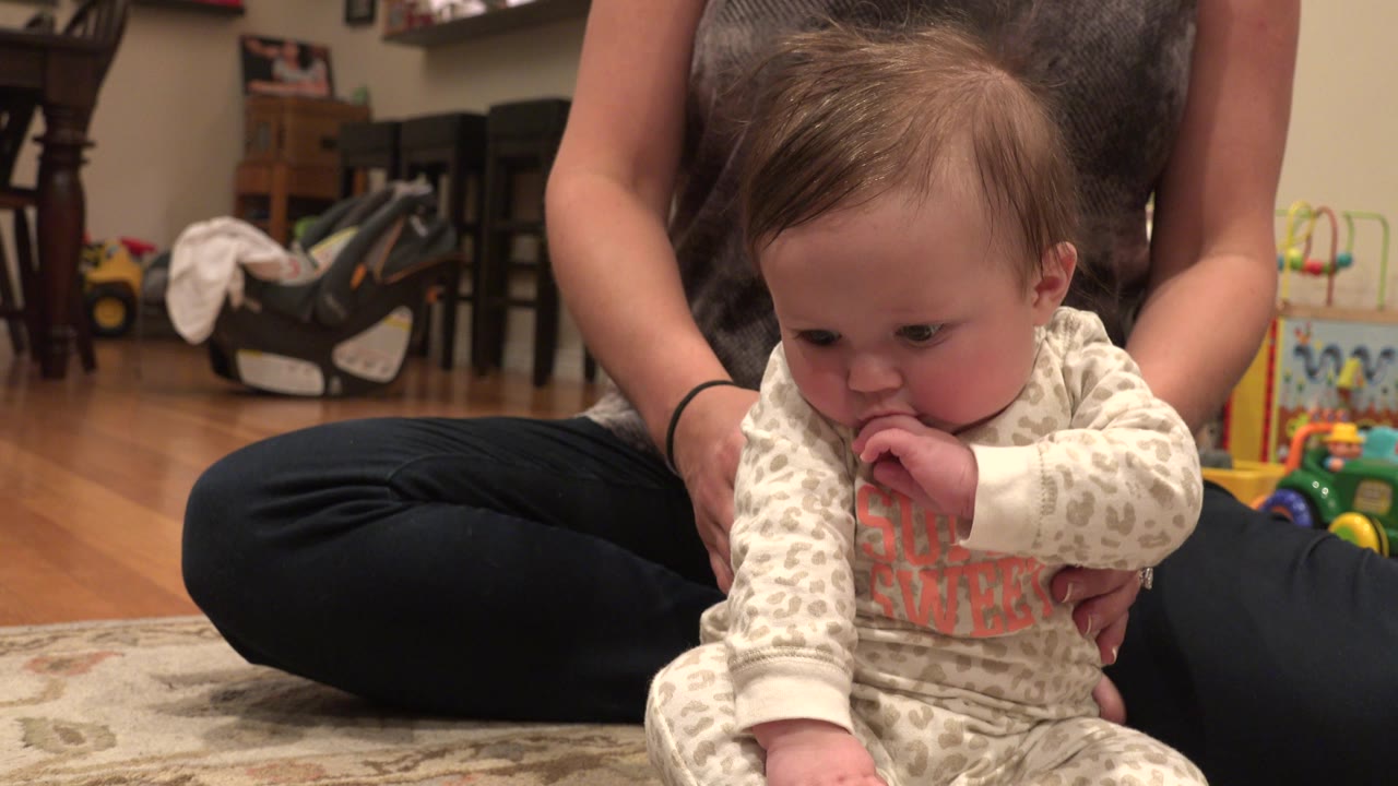 Baby turns slightly right with a hand in the mouth while seated in front of a woman.