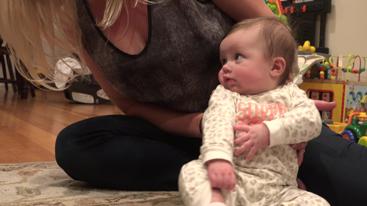 Baby sitting on a rug while a woman supports them from behind.