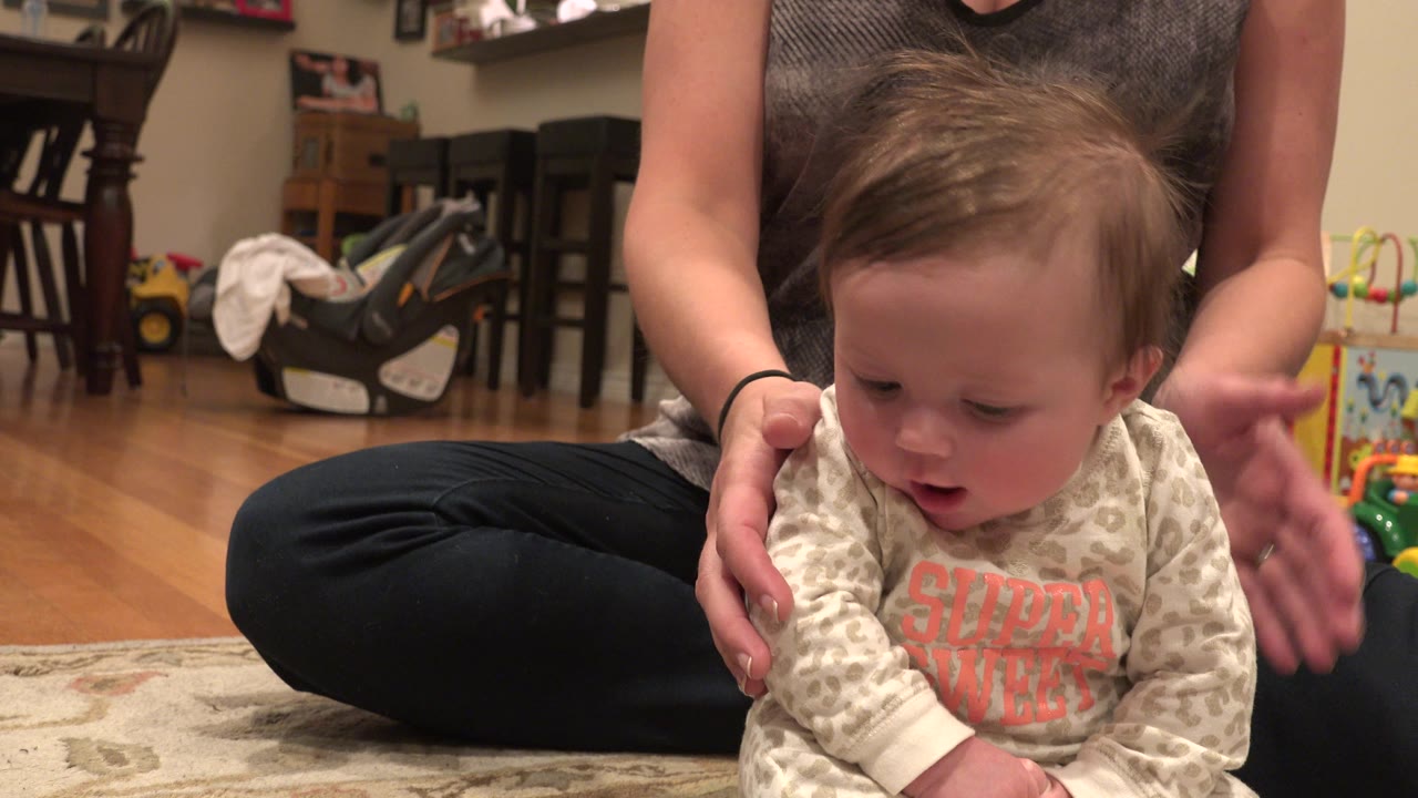 Baby sitting on a rug while a woman supports them from behind.
