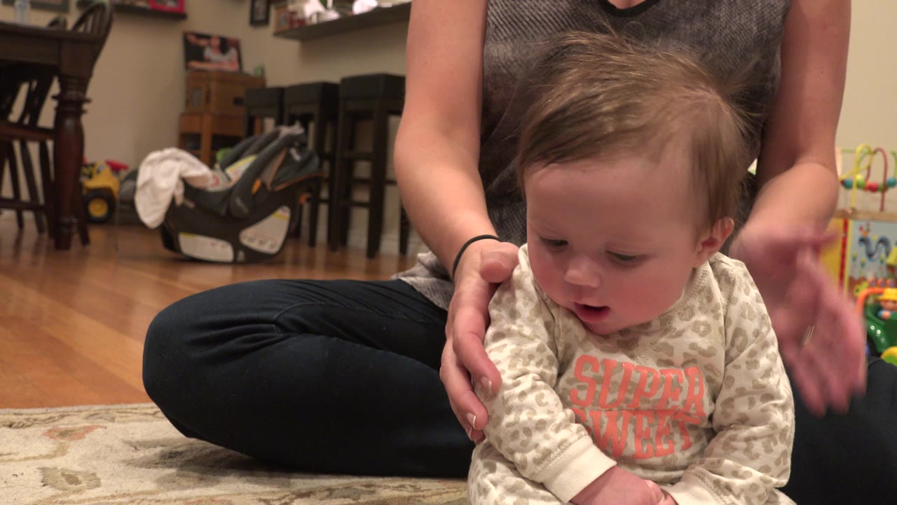 One second in, the baby remains seated in front of the woman's crossed legs, with the caregiver's hand at the baby's side and toys visible behind them in the Westminster home.