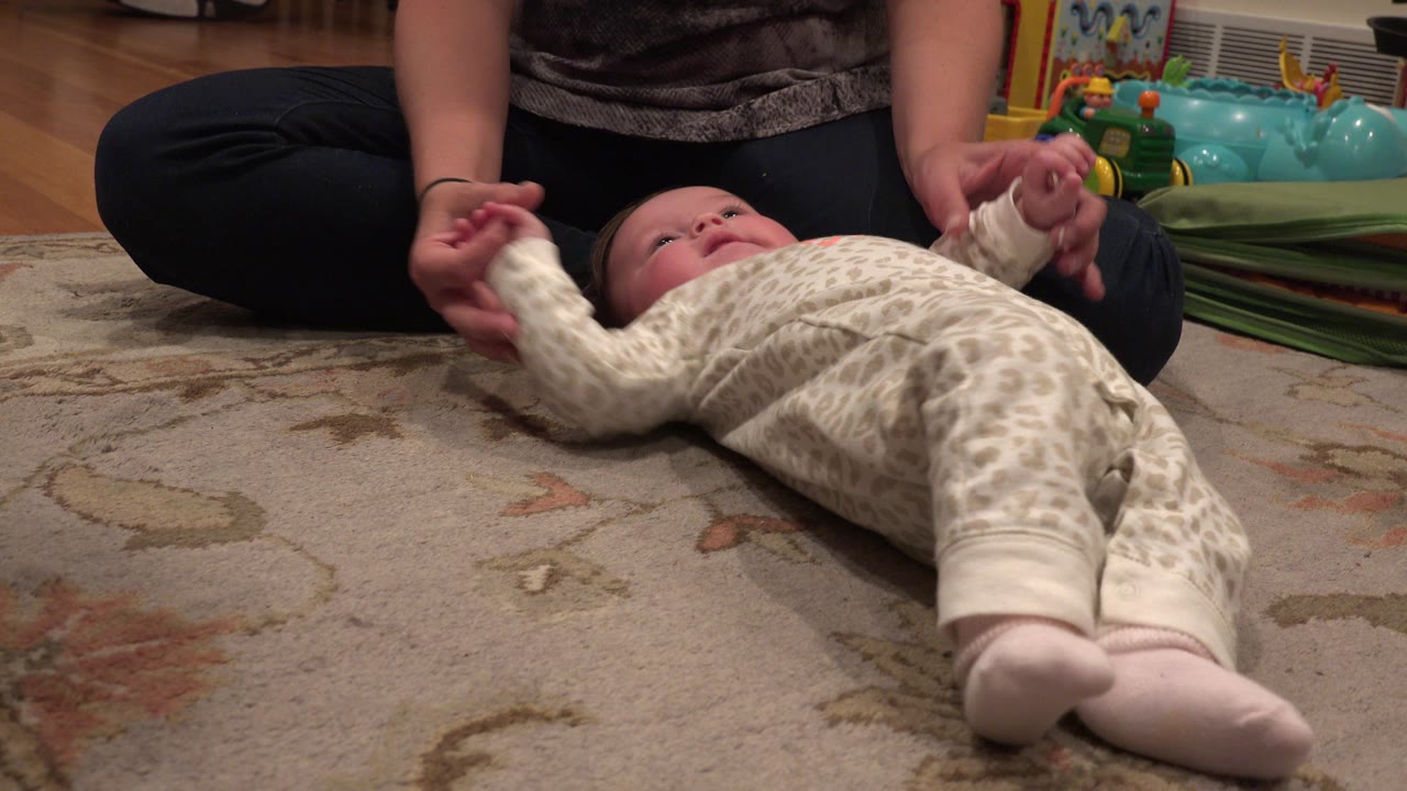 Baby lying on a rug while a seated mother holds both hands.