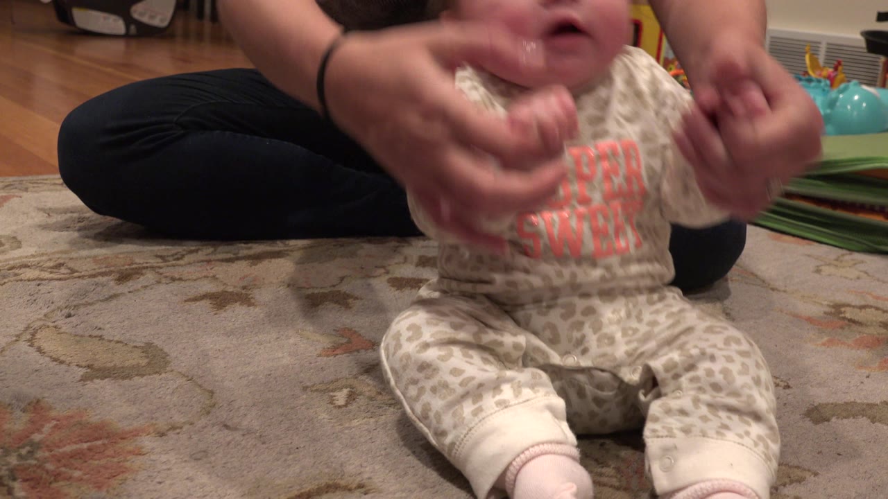 Infant seated on a rug while an adult steadies their arms.