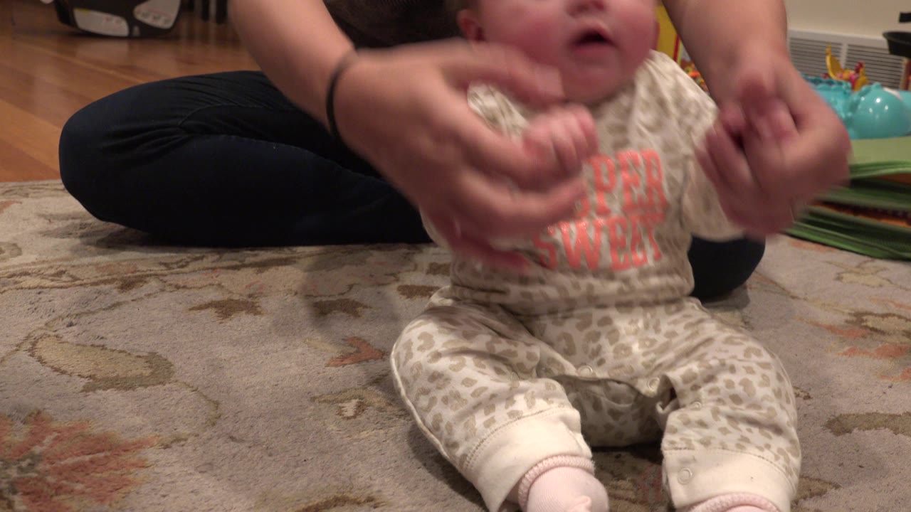 At 0:01 in Westminster, Colorado, the infant remains seated close to the lens while the adult steadies both arms, with toys and a play mat behind them.