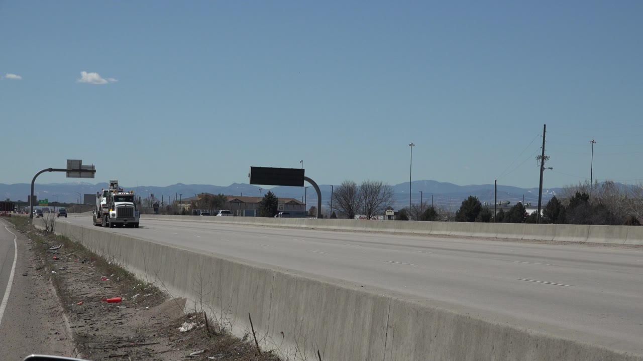 The large white truck is closer on the Denver roadway, with cars spread across the lanes, a concrete median barrier, roadside signs, bare trees, distant mountains, and no visible people.