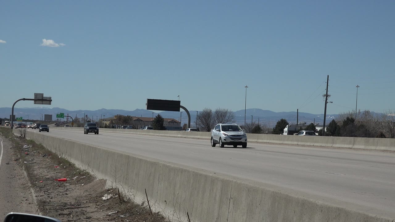 A Denver, Colorado road scene shows several cars traveling under a clear blue sky, with a concrete barrier in the foreground, overhead signs, distant mountains, and no visible people.