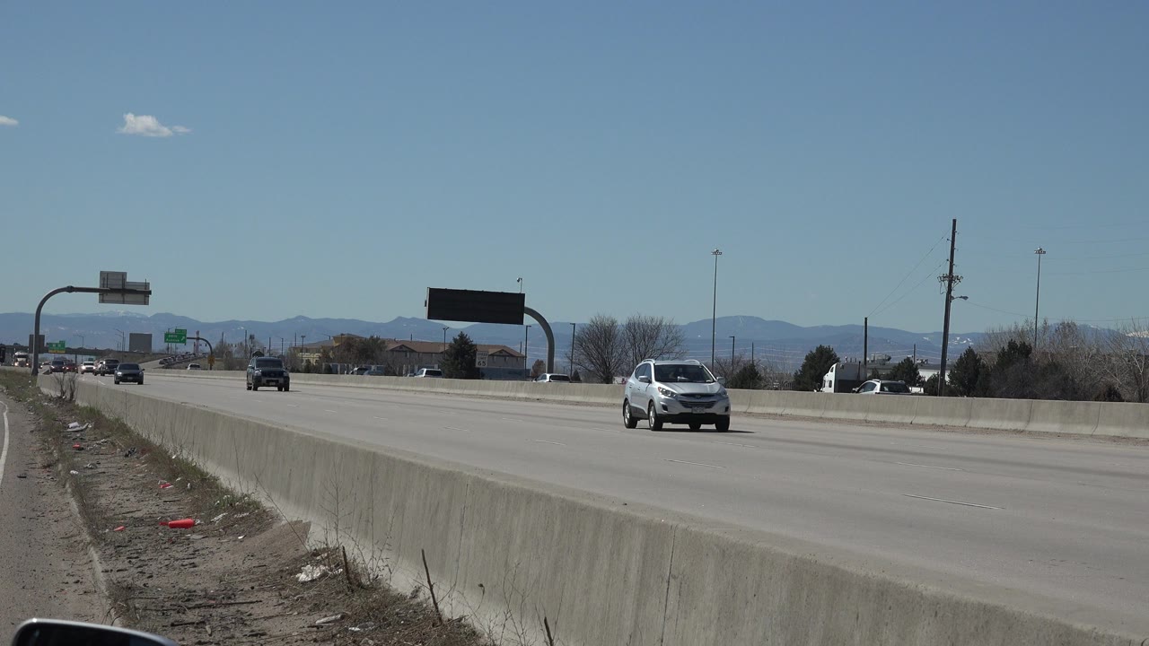 A white car moves closer on the Denver roadway while other vehicles remain farther back, with the barrier, road signs, utility poles, mountains, and no visible people in frame.