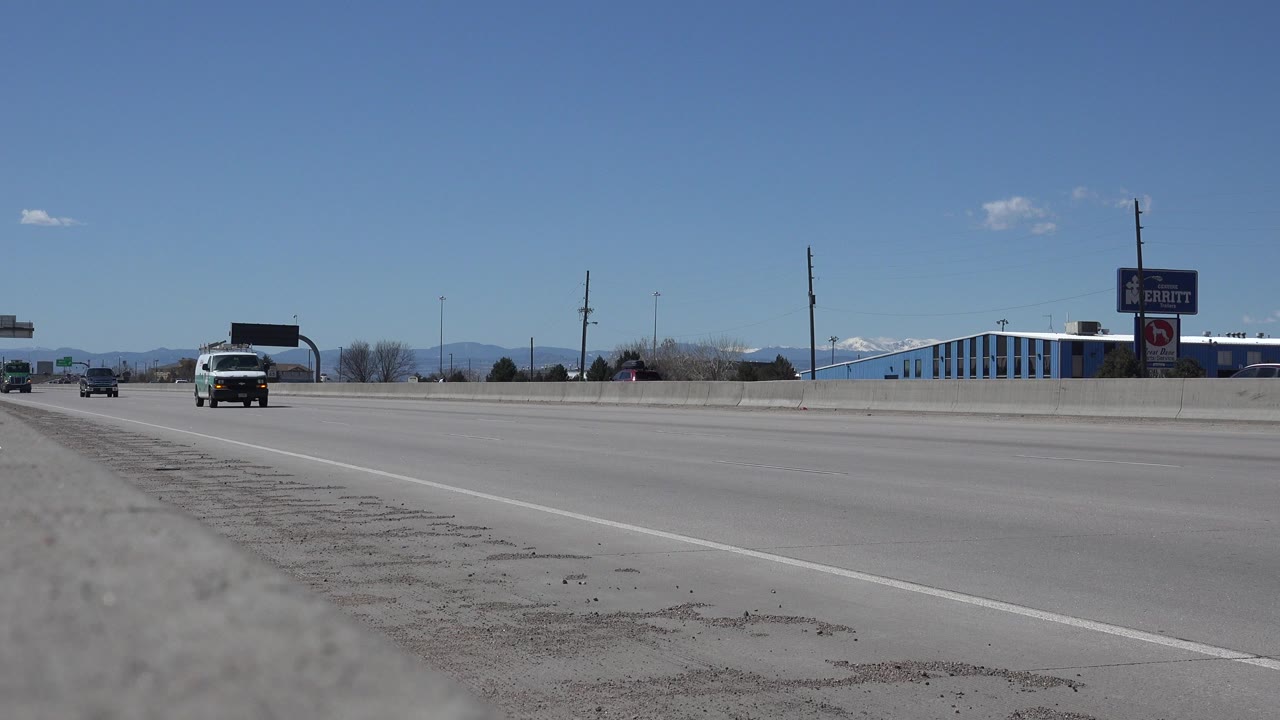 Denver, Colorado highway view at the start of the clip, with a white van and small cars in the distance, roadside signs, low buildings, mountains, and no visible people.