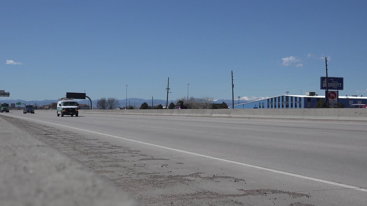Denver, Colorado roadway with the white van slightly closer, small cars farther back, a blue-and-white roadside building, commercial signs, mountains, and no visible people.