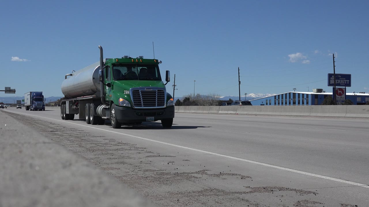 The green tanker truck is closer to the ground-level camera on the Denver highway, with open lanes, commercial roadside signs, and no visible people.