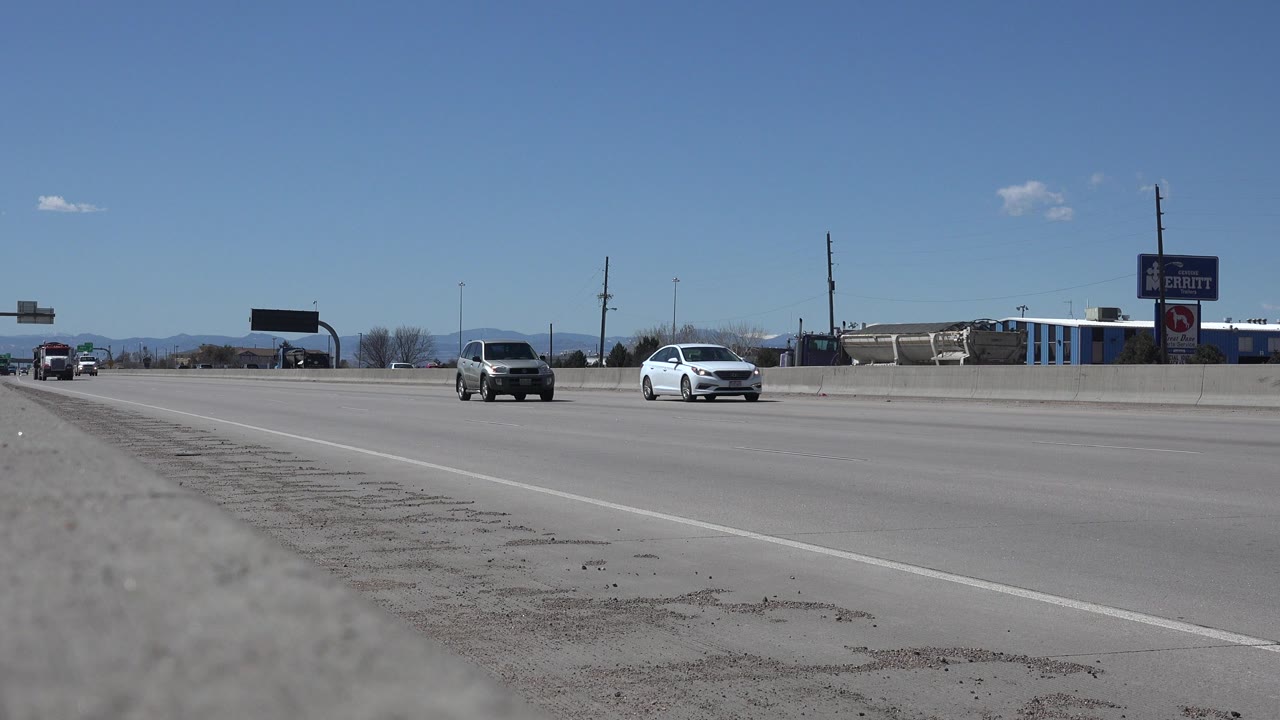 Low roadside view of Denver highway lanes with two cars approaching, gravel shoulder in the foreground, distant mountains, signs, and no visible people.