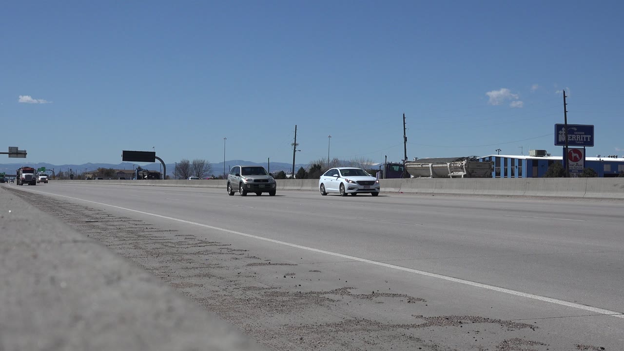 Denver roadway frame with a dark car and a white car traveling through open lanes, with roadside buildings, poles, signs, mountains, and no visible people.