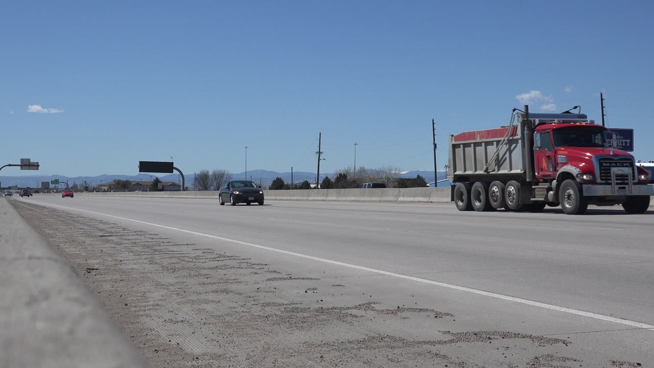 The red dump truck is farther across the Denver highway frame, with a dark car behind it, open lanes, a gravel shoulder, distant mountains, and no visible people.