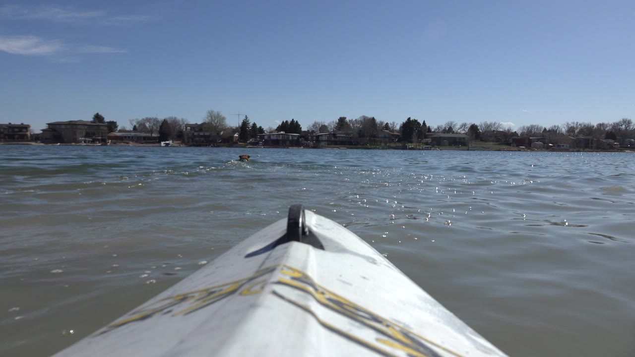 A white pointed watercraft bow enters the lower frame on Hidden Lake in Colorado, with rippled murky water, a distant shoreline of buildings and trees, and no visible people.