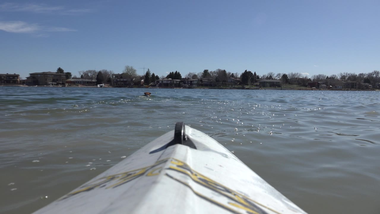 The white bow remains centered over choppy Hidden Lake water, with sun glints on the surface, low buildings along the horizon, and no visible people.