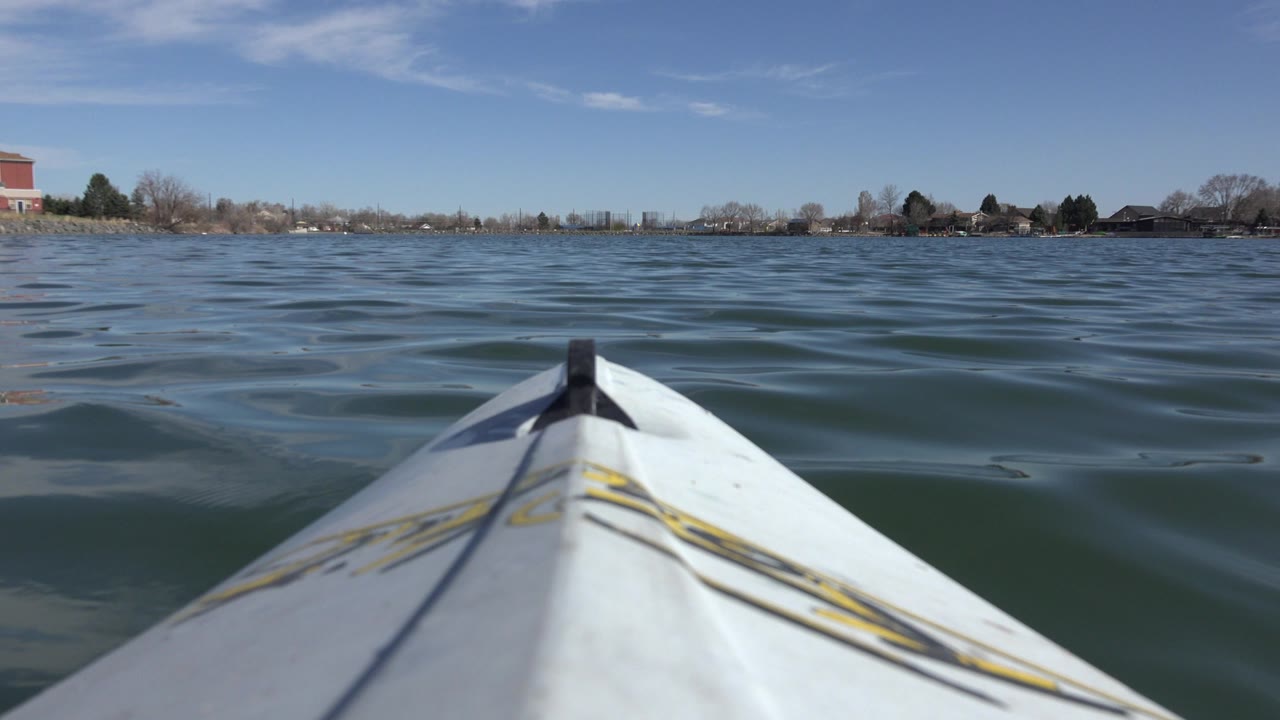 White kayak bow points across rippled water on Hidden Lake, Colorado, with a distant shoreline, houses, bare trees, blue sky, and no visible people.