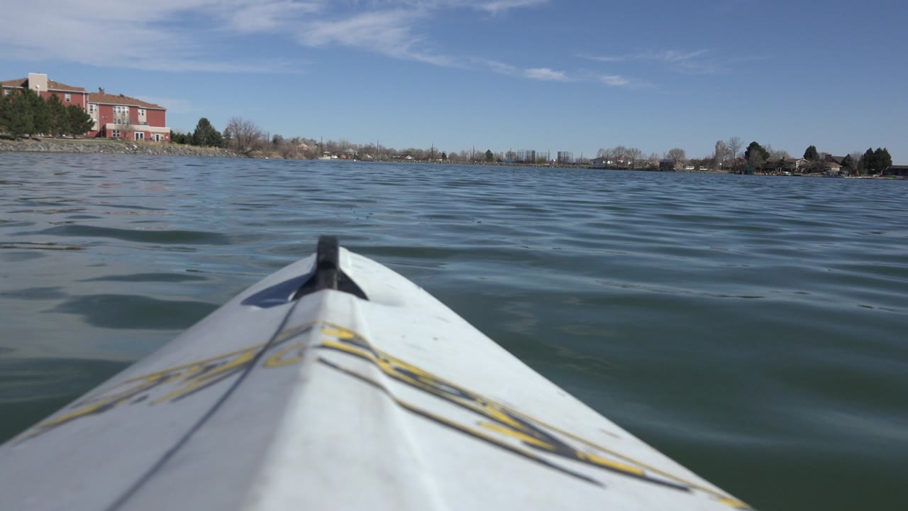 Centered canoe bow on Hidden Lake, Colorado, with small wave bands crossing the water, a red building at left on shore, bare trees, and no visible people.