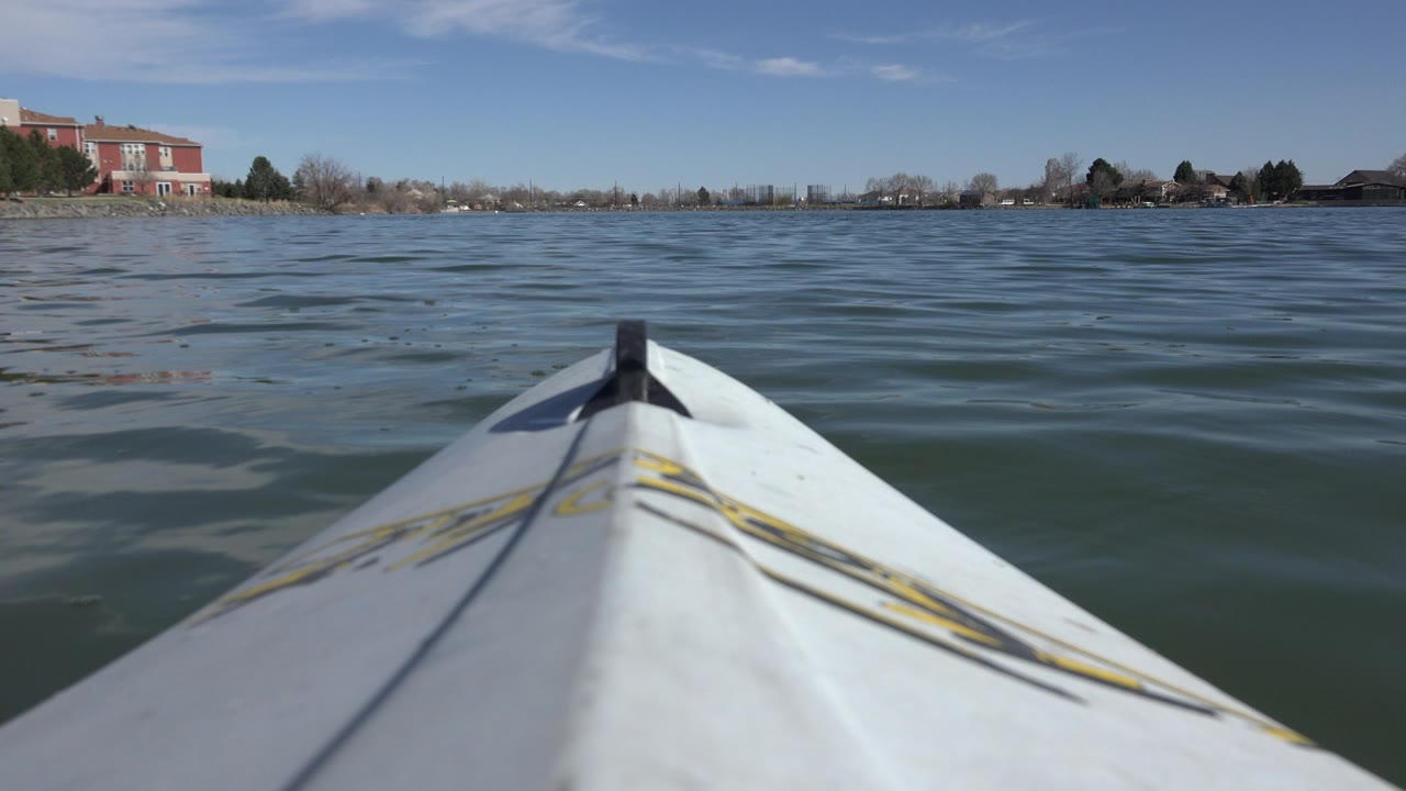 At 0:00, a white boat bow fills the lower frame on Hidden Lake, Colorado, with rippled water, trees, and red brick buildings in the distance, with no visible people.