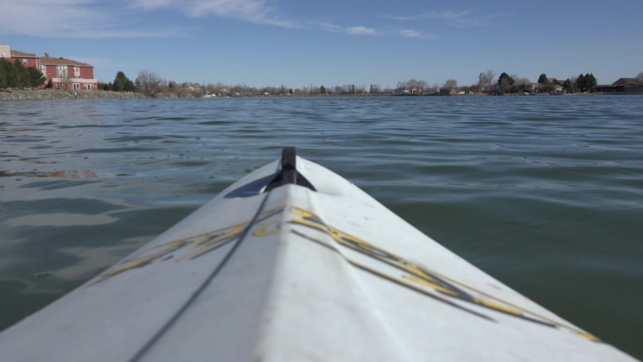 At 0:01, the boat bow remains centered on the water, with a low Hidden Lake shoreline, bare trees, evergreen shrubs, and red buildings beyond, with no visible people.