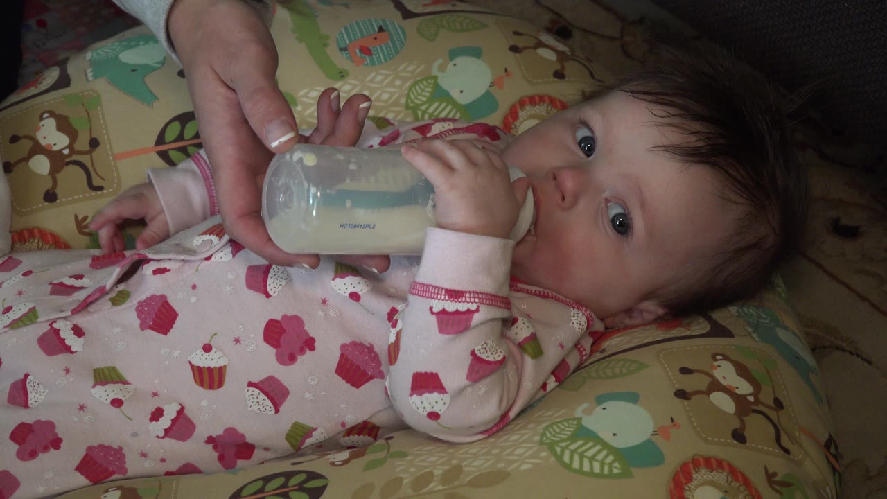 Baby lying on patterned bedding while drinking from a bottle held by an adult hand.