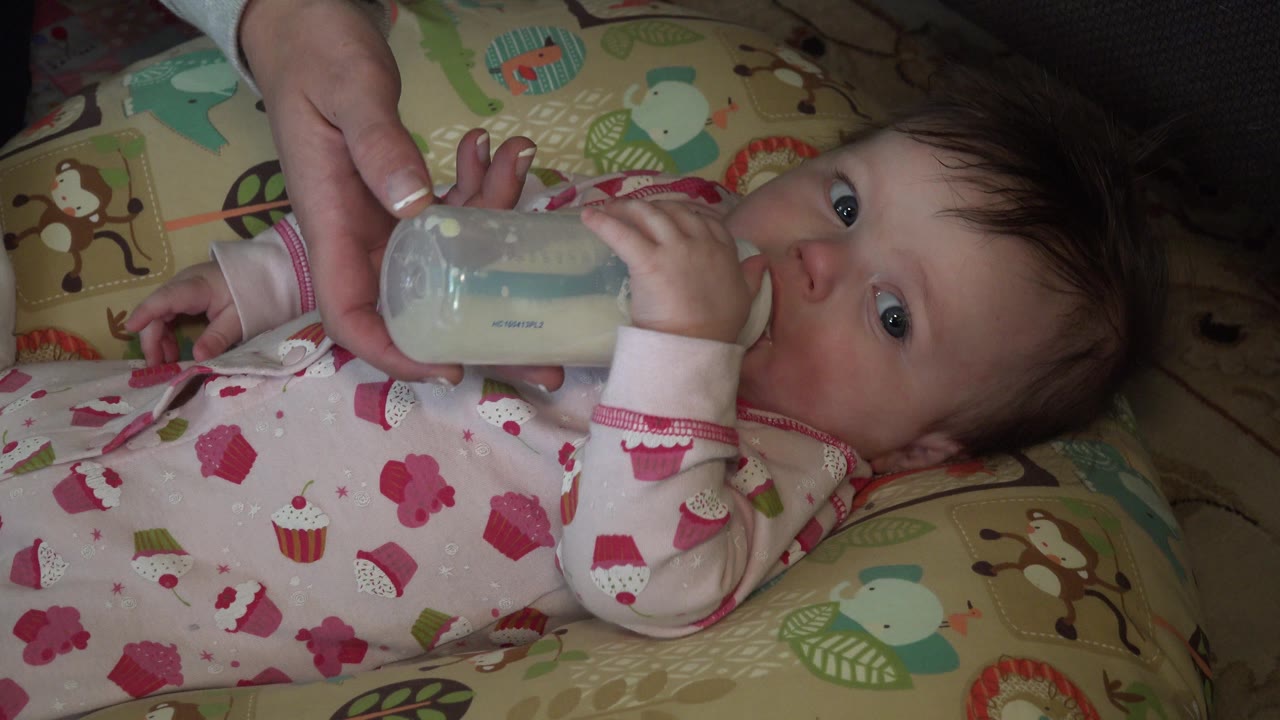 At 0:01 in Hidden Lake, Colorado, the baby's eyes are open toward the camera area as an adult hand steadies the bottle over patterned bedding.