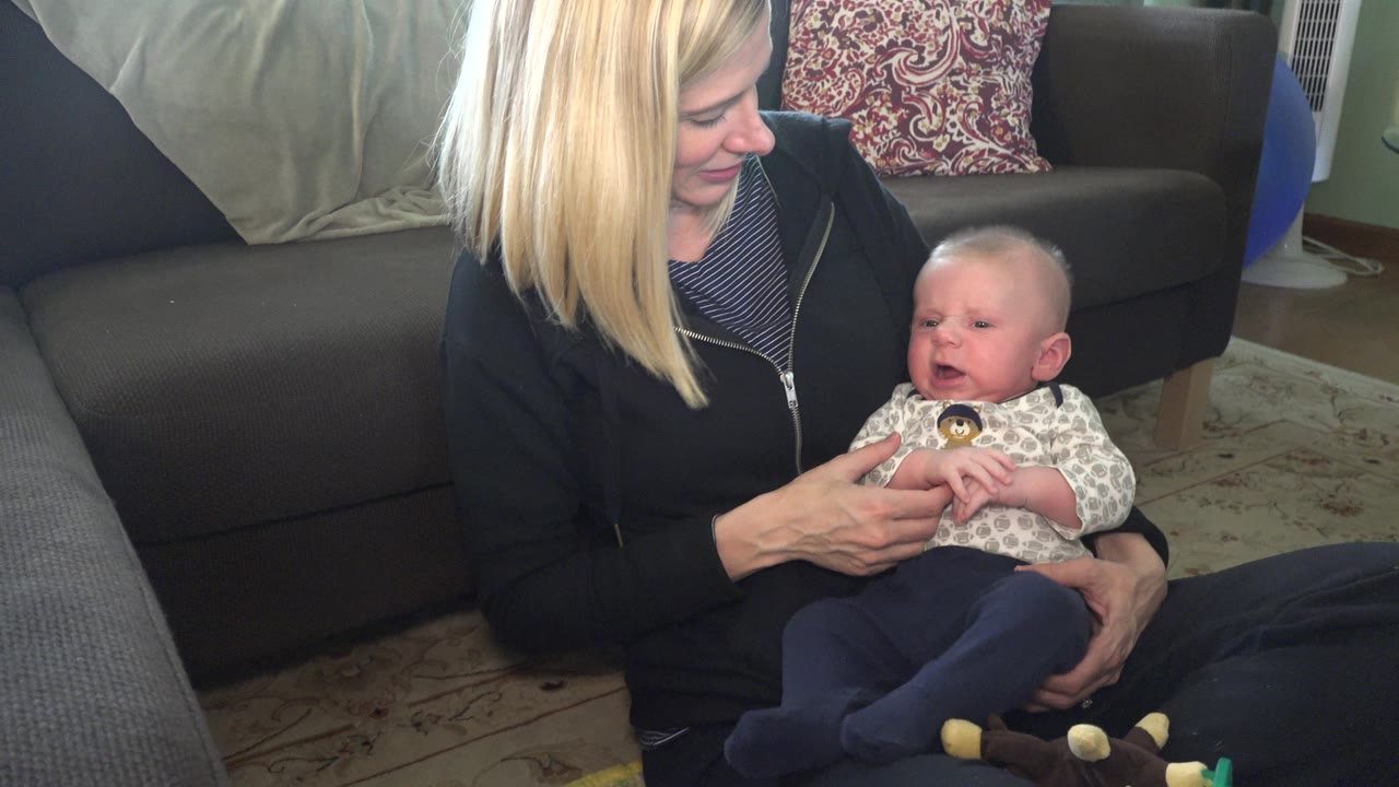 In Hidden Lake, Colorado, the mother keeps her hands around the baby as the infant's mouth opens slightly, with couch cushions and a patterned pillow behind them.